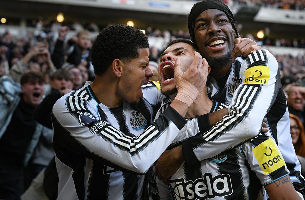 Several male Newcastle United players in black and white striped jerseys embrace in celebration during a football match. The background shows cheering fans in a crowded stadium.