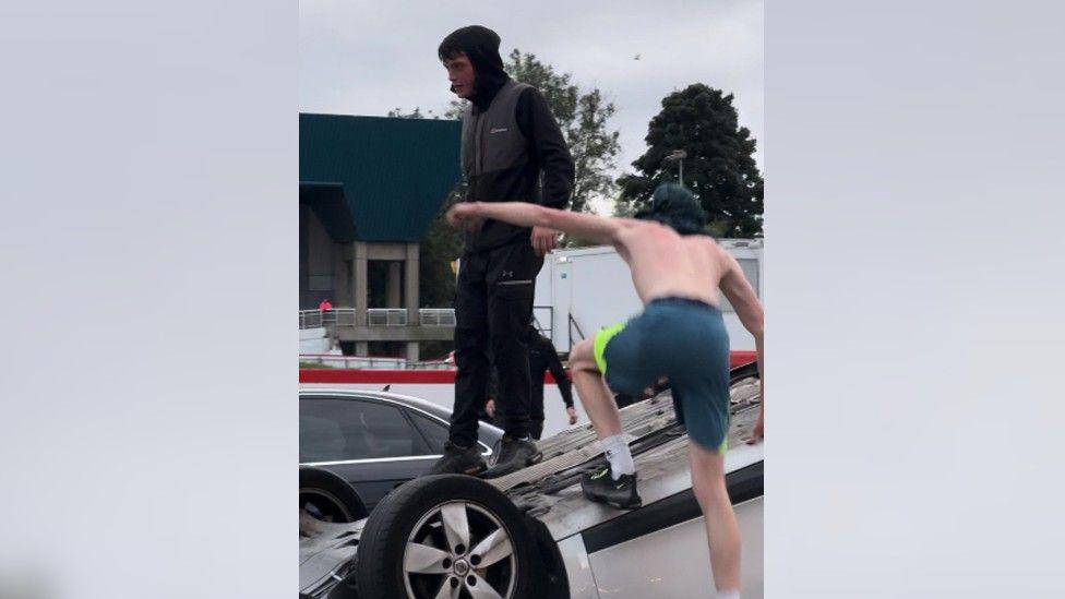 A young man dressed in black stands on top of an upturned car, while another shirtless boy with his back to the camera clambers up to join him.