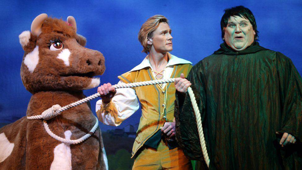 A man wearing a black cloak and a man wearing a yellow and white outfit on stage at the pantomime. They are using rope to pull along a fluffy cow, which has an actor inside.