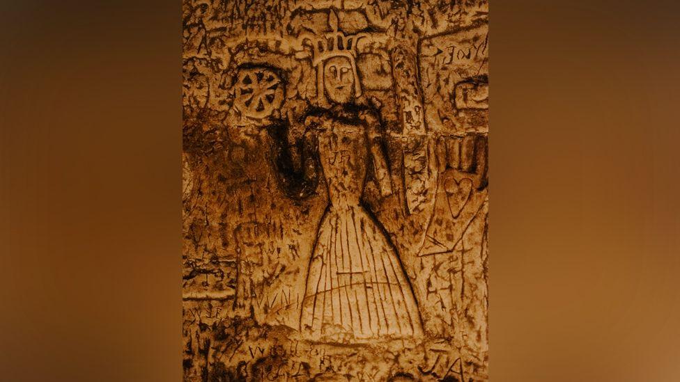 A close up of the carving believed to be St Katherine who wears a dress and a crown on her head, while to her left she holds an eight spoked wheel. Other markings are etched into the wall around her.