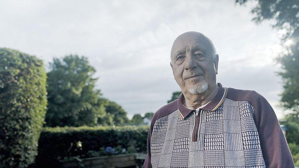 John Morris, with a white beard, stands in a garden and looks wryly at the camera. 