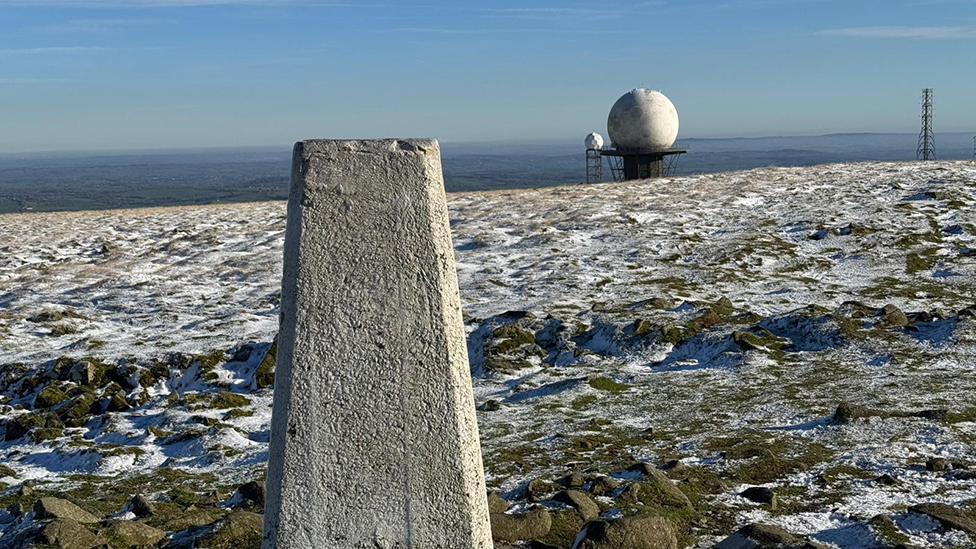 The summit of a snow-covered Clee Hill, in Shropshire. A trig point is in the foreground, with white radar domes behind