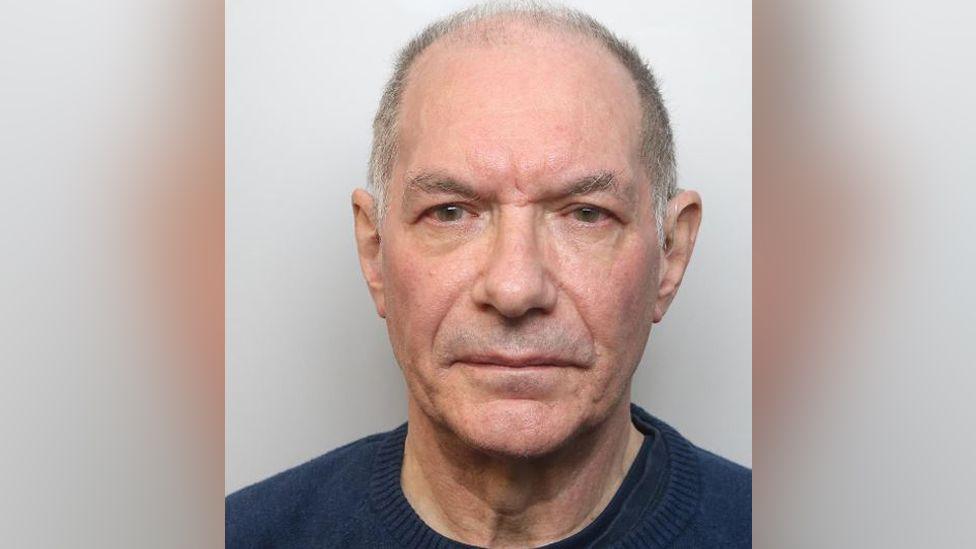 Police mugshot of Robert Gould. He is balding with close-cropped grey hair. He is clean-shaven and wearing a blue crew-neck sweater over two darker T-shirts. He is looking directly at the camera with a neutral expression.