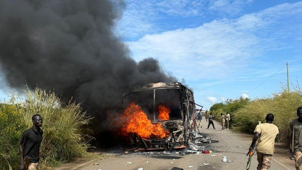 A bus on fire on a road - black smoke rises above it.