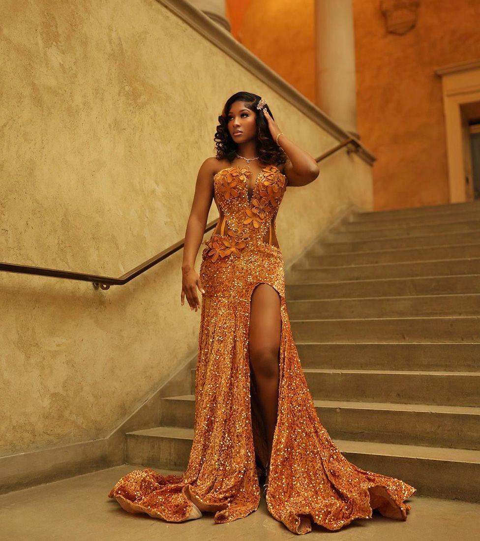 Anita Konneh wearing an ochre, sequined dress with a split by her leg. she is standing on a staircase - Greco-Roman columns can be seen behind her.