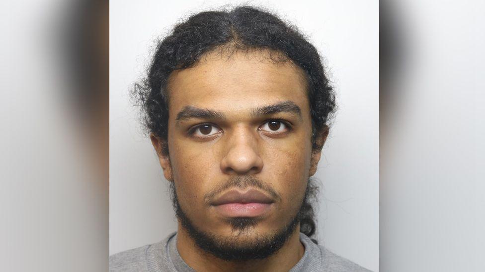 Jayden Gubetini mugshot pictured looking straight at the camera, not smiling. He has a goatee beard and hair tied back in a ponytail, brown eyes and wearing a grey sweater.