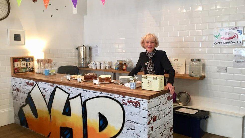 Denise Mackay with blonde hair wearing a dark top and a scarf, standing behind a white brick cafe counter with cakes on top and the initials "JHP" painted on the front