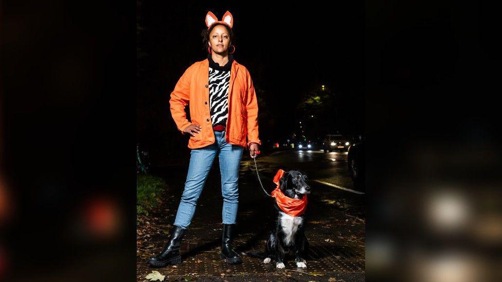 A woman wearing a pair of fox ears and an orange jacket, standing on a dark pavement with one hand on her hip and staring unsmiling at the camera. She has a black and white dog on a lead which is wearing an orange neckerchief.