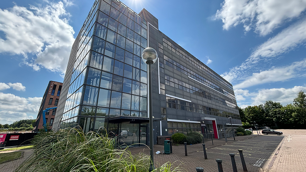 A modern-looking glass and grey office block surrounded by car parking.