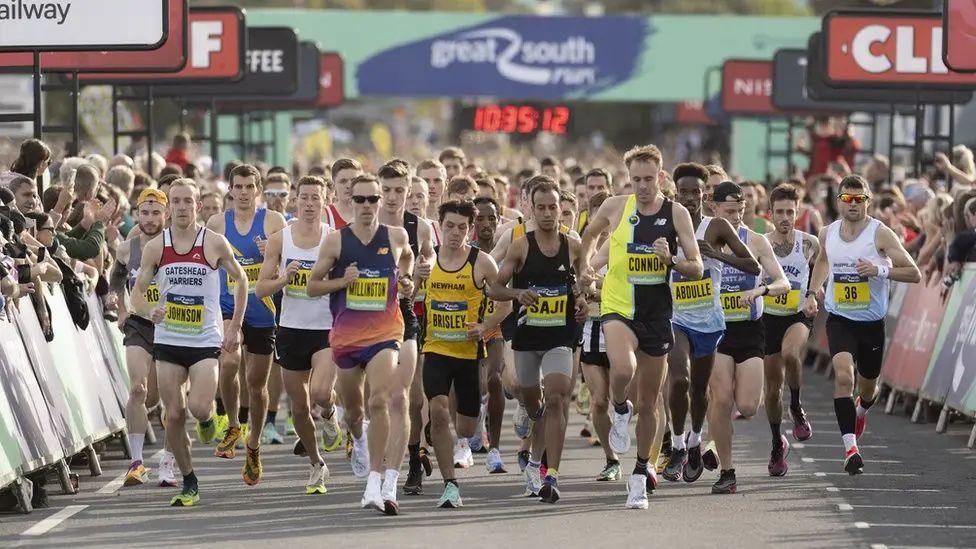 Leading runners in the 2022 Great South Run run along a road towards the camera. Crowds of cheering spectators stand behind advertising hoardings on either side