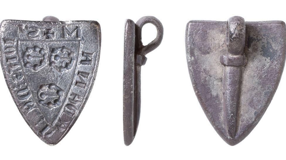 Three views of a silver medieval seal matrix. The view on the left shows the front of the shield-shaped seal. It has writing around its edge and in its middle are three brooch-shape designs, two above the third. The middle view is side on, showing its edge and its suspension view. The right view is plain and shows the long tail of the suspension loop attached to the back.