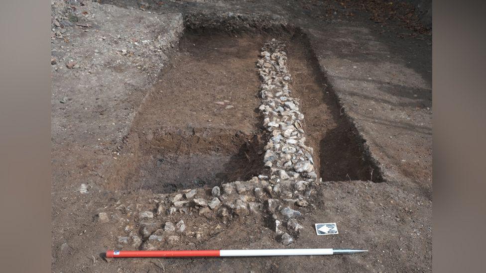 A rectangle shaped trough dug into the soil lined with stones, and a red/white measuring instrument lying at the foot of it.