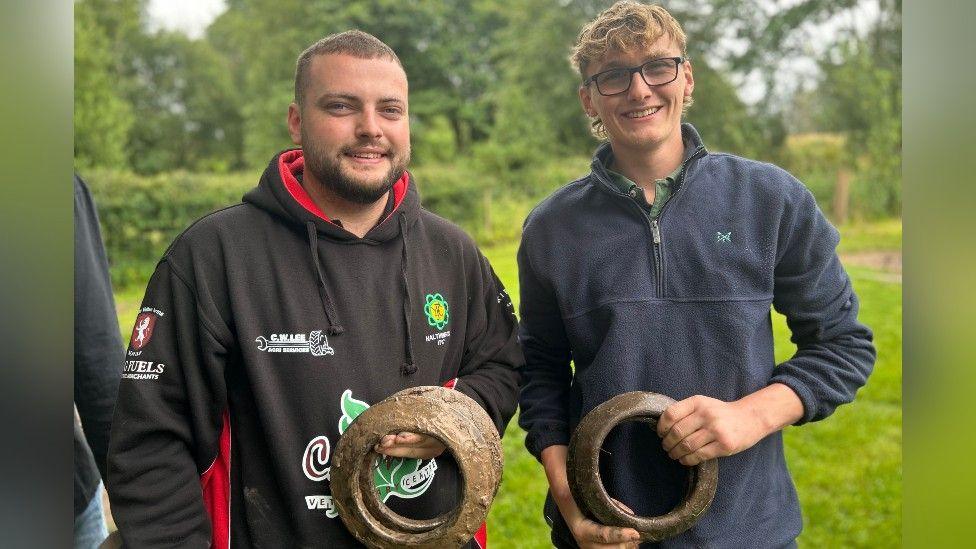 Cameron Robbie and Johnnie Redden are smiling as they each hold a quoit. Cameron has a short brown hair and blue eyes and is dressed in a black hoodie. Johnnie has wavy blonde hair and is wearing glasses and a navy blue half-zip jumper with a green shirt.