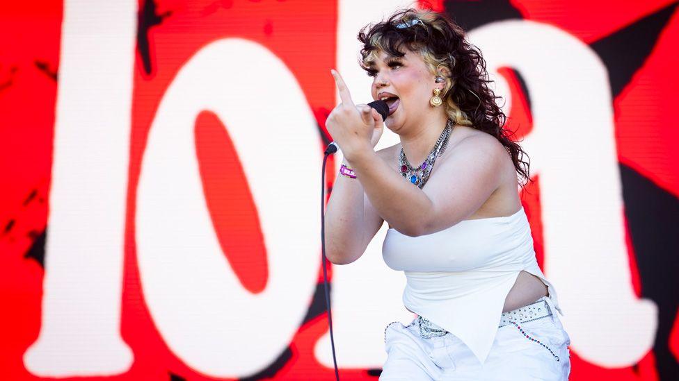 Lola Young performing on stage with a finger raised, in front of a red and white backdrop saying "Lola"