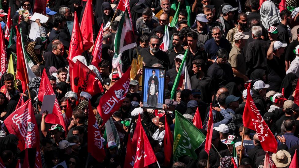 Məşhəddə İranın Ali Lideri Əli Xameneinin dəfn mərasimi üçün toplaşan yas tutanlar. Çoxları ərəb yazılı qırmızı bayraqlar daşıyır, ortada oturmuş Xameneinin şəklini tutan bir kişi, ondan yuxarda isə oğlu Məcəddə durur.