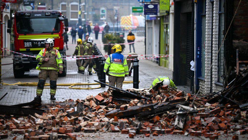 Firefighters and fire engine in street with piles of burnt timber and rubble strewn across from collapsed building.