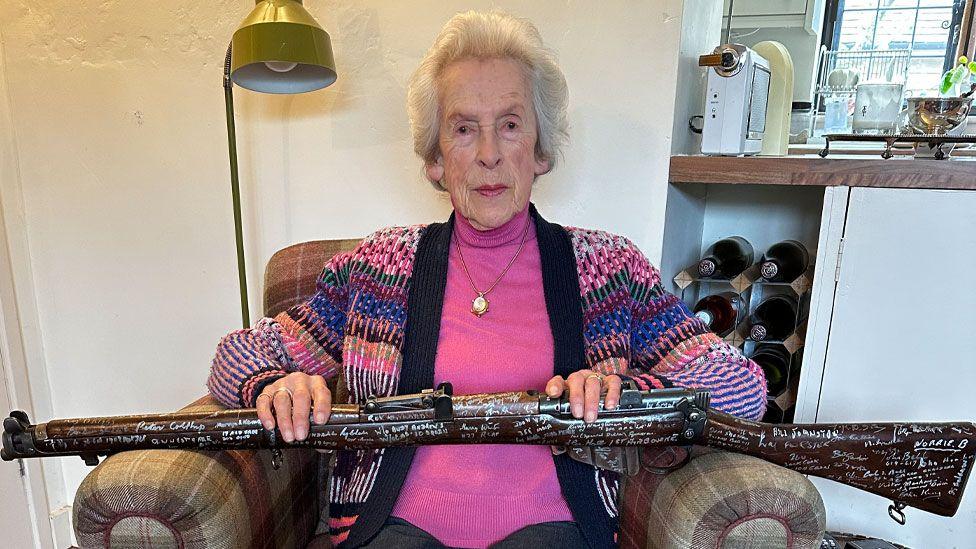 Joyce Wilding with grey short hair wearing a pink roll-neck under a multicoloured cardigan sitting on a plaid arm chair holding a Lee-Enfield rifle covered in signatures