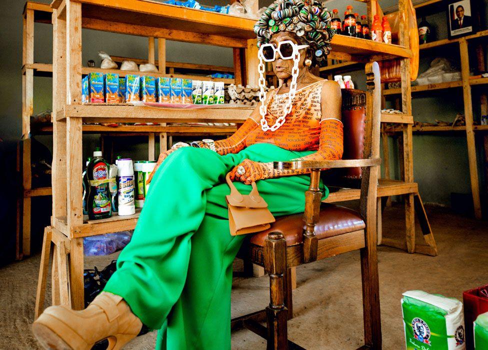 Margret Chola sitting on a wooden chair in what looks like a rural store with produce on shelves. She is wearing a hat shaped like an afro made from bottle tops, massive white-framed sunglasses with a chunky white chain, green flairs, an orange sleeveless top with matching long gloves, big high-heel camel-coloured platforms and clutching a small handback of the same colour