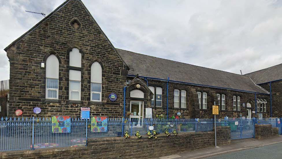 Street view image of Sharneyford Primary School in Bacup, a single storey  black stone building with blue metal railings and bright artworks on the front