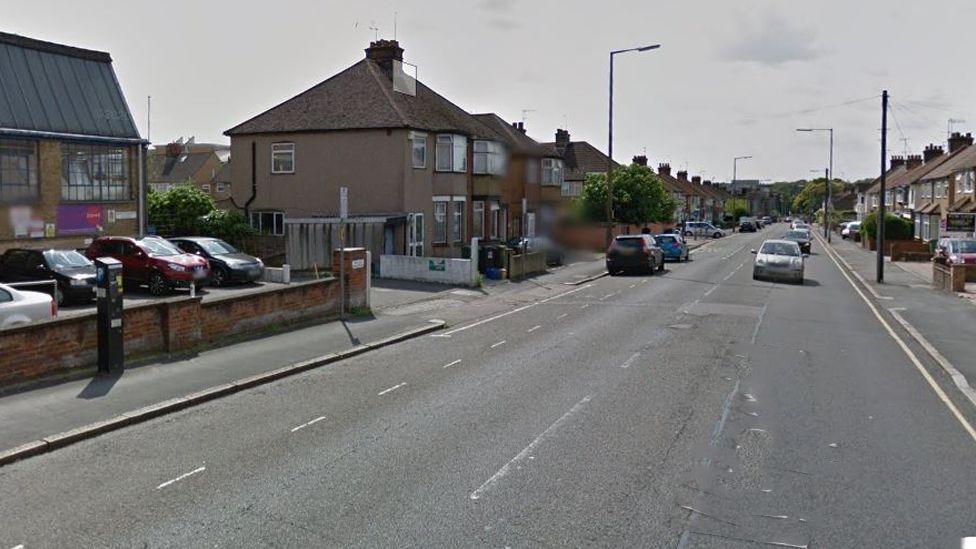 Whippendell Road, with some traffic coming towards the camera. There is a parking machine on the left-hand pavement. Houses line both sides of the street. There are dotted lines on the left side marking parking spaces, and a single yellow line on the right.