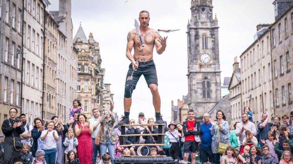 A man juggling three large knives balances on a a precarious contraption of bricks, planks and tubes. He is topless and wearing blue shorts and has tattoos. 