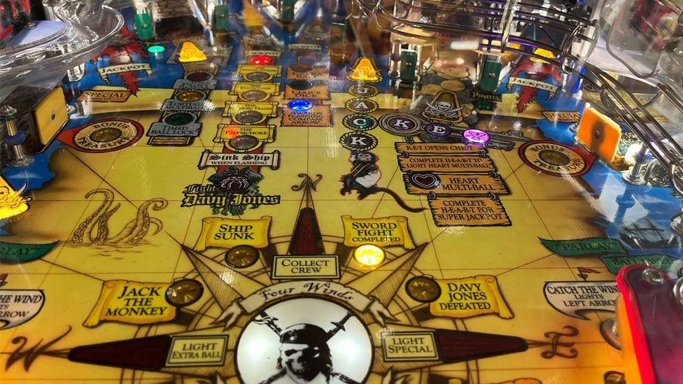 A close-up of a very colourful Pirates of the Caribbean pinball machine table, with a yellow background, writing and lights glowing