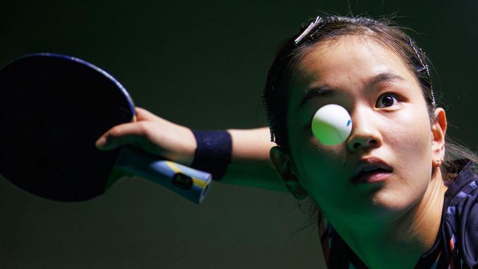 A close-up of a table tennis player holding a paddle in a ready position, with the arm extended and wristband visible. The ball is seen in mid-flight over the player's right eye.