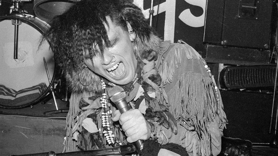 Black and white photograph of Pete Burns on stage crouched on stage singing into microphone wearing tasselled jacket