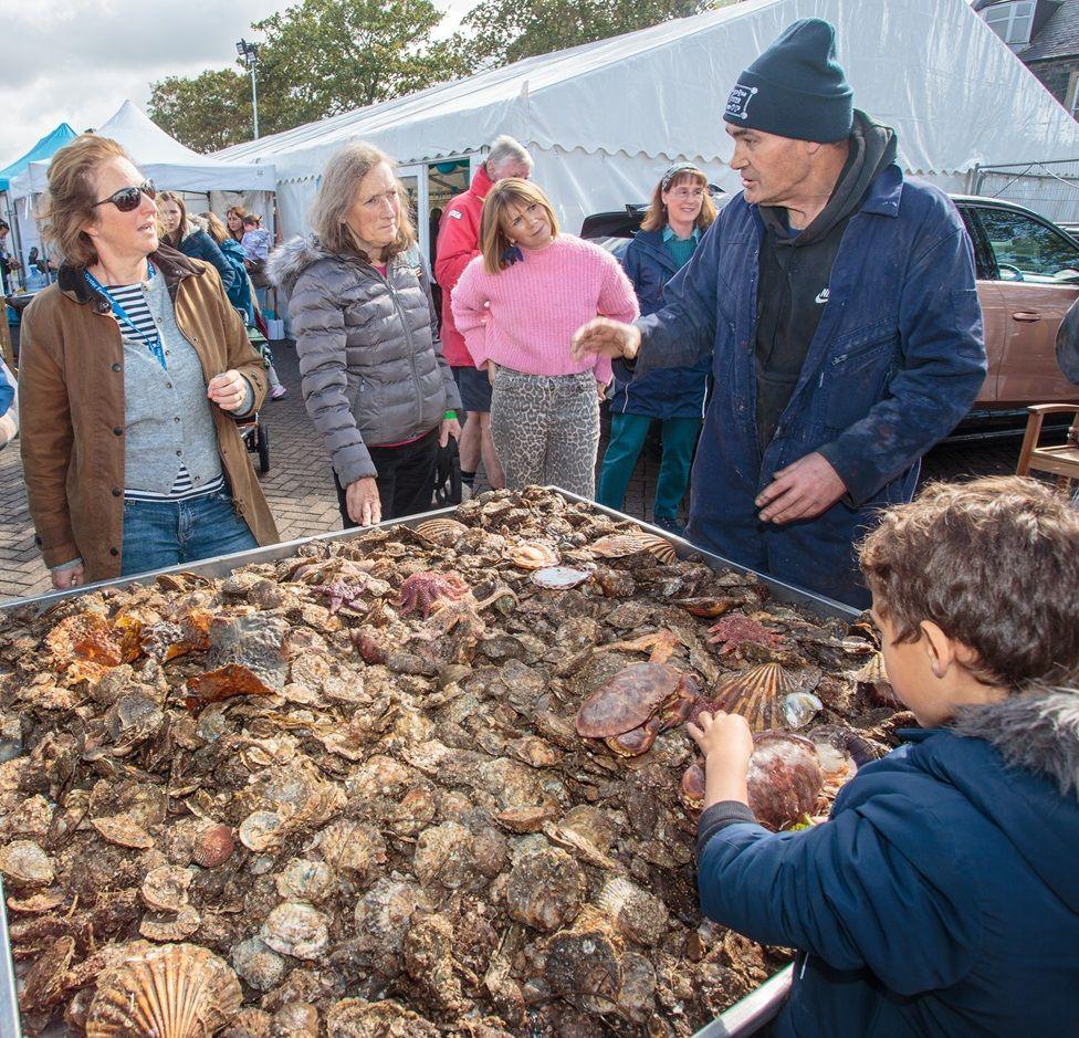 A man in a woolly had and blue overalls talks to a group of people while a child picks up a shell from a large tray of shells