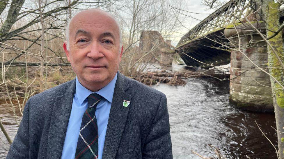 Marc Macrae standing at the Spey Viaduct, the damaged section is visible in the distance, in the river.