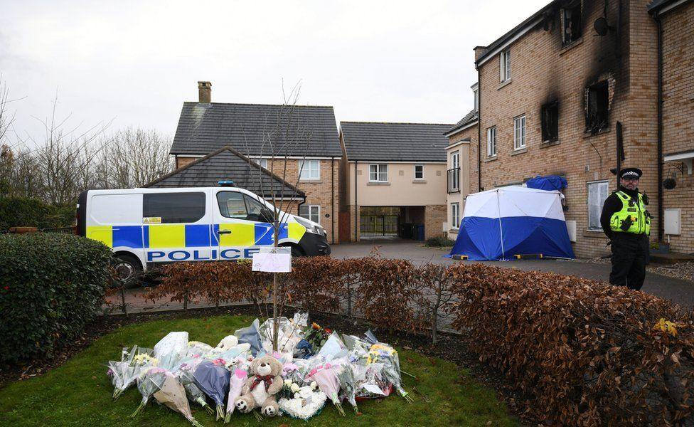 A terraced modern three-storey house with fire damage around the windows and a police van and officer, plus piles of flowers, Eynesbury, Cambridgeshire