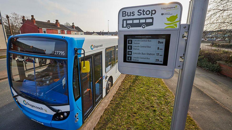 View of one of the new electronic bus stops mounted on a metal pole with a list of bus numbers on the screen a blue and white Stagecoach bus is parked by the stop