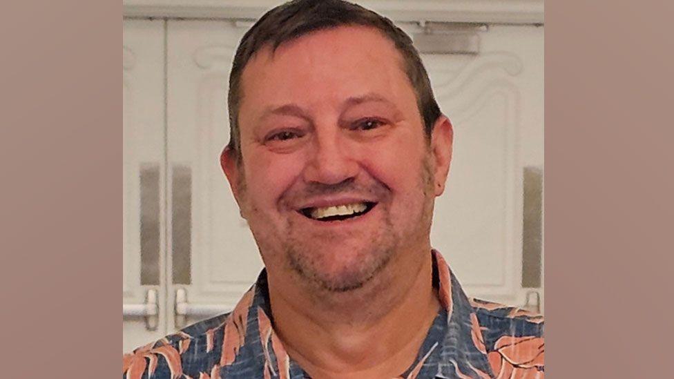 Anthony Liddell smiling broadly at the camera. He has short light brown hair and is wearing a blue shirt with an orange floral pattern which is open at the neck.