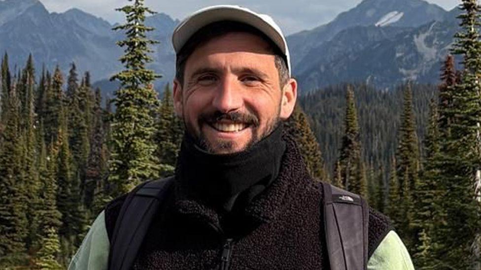 Oliver Gooch standing smiling in front of trees and mountains smiling. He has dark hair and a beard and is wearing a white baseball cap and black fleece and scarf