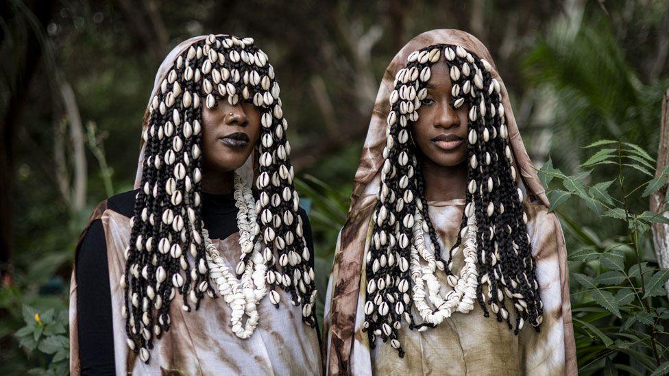 Two women stand near trees. Their clothes are visually striking and their facial expressions are calm. In Dakar, Senegal - Saturday 7 December 2024