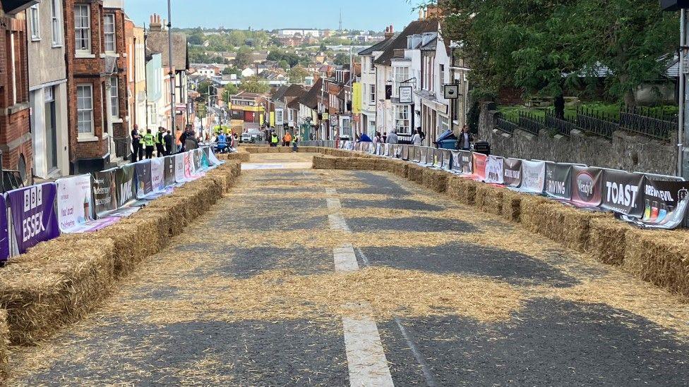 Hay bales and silver fences are lined up on either sides of a road. On the silver fences are sponsorship banners. Buildings are on the sides of the road and people are standing behind the fences.