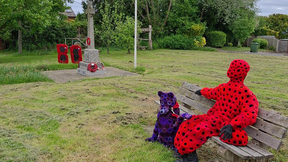 A VE Day memorial featuring a figure covered in poppies sitting on a bench with a figure of a dog next to him. In the background there is a wreath in figure of 80 made from poppies next to a war memorial, with grass in the foreground