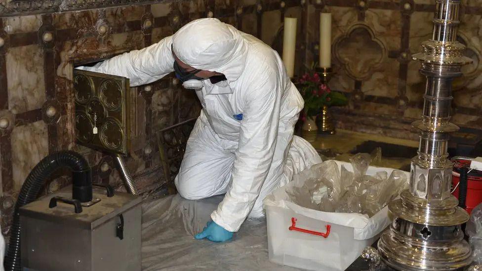 A person in a white protective suit on their hands and knees in a church. To the person's left is a white plastic box filled with clear plastic bags. the person is reaching into a small square door in the church's wall