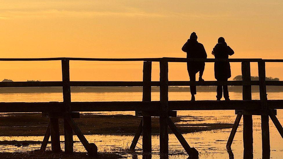 Two people look out across an expanse of water while standing on an old wooden pier. They are silhouetted against a pale orange sky at sunset.