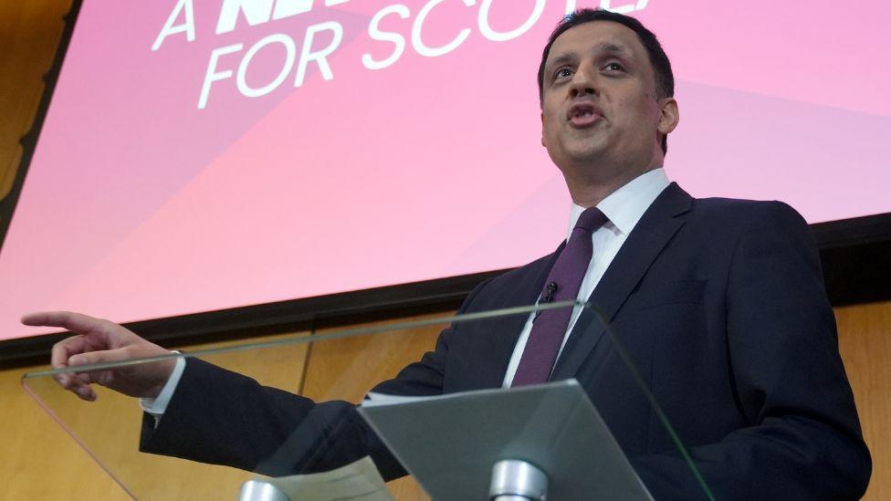 Anas Sarwar with dark hair, wearing a dark suit, white shirt and purple tie, points to his right while speaking on stage. He is photographed from a low angle, standing behind a podium and in front of a red Labour sign. 