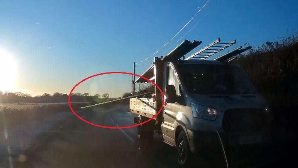 A white van with ladders on the roof. A board coming from the back of the van is circled in red.