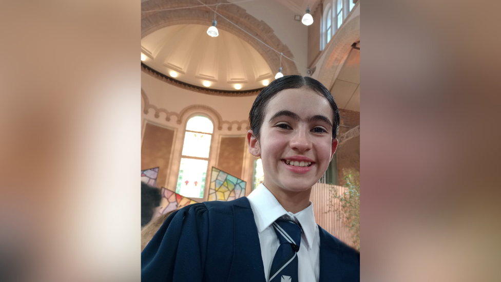Zoe. She has dark hair tied back, wearing a navy school uniform, white shirt and navy striped tie. She is standing in a church.