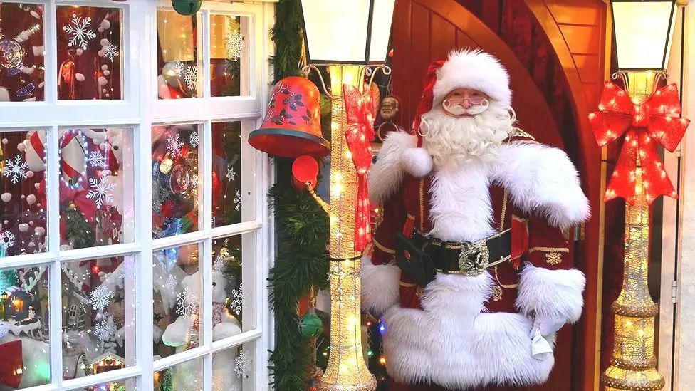 Santa in a red suit and white trim stands by a window with festive decorations