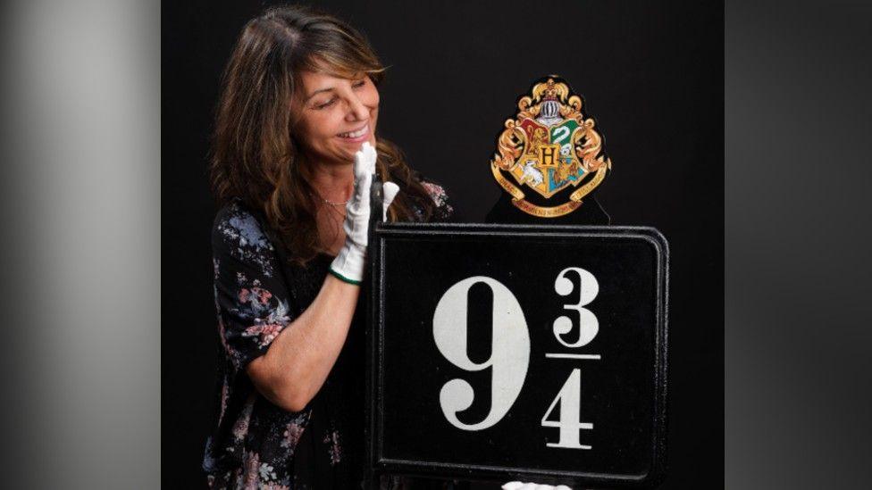 A woman with brown hair standing next to a sign which is from the Harry Potter original movies which is for Platform 9 3/4. 