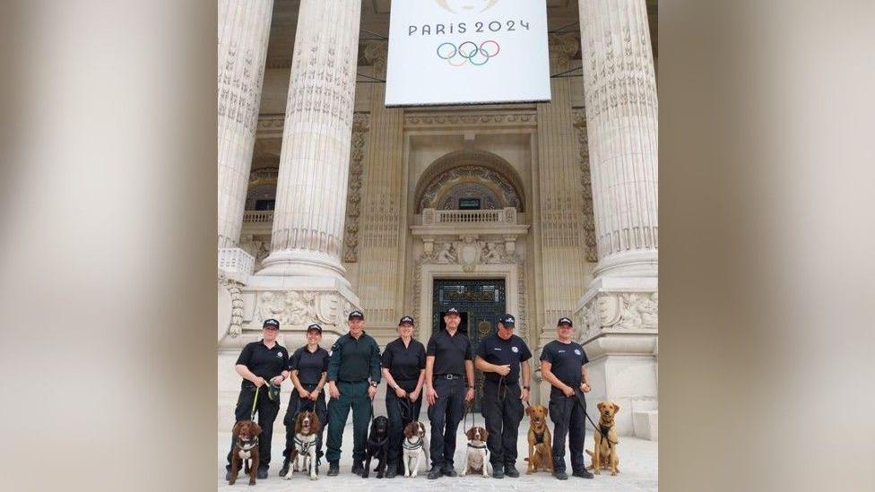 A row of uniformed police officers with their dogs outside the embassy, with white columns and a Paris 2024 Olympic poster