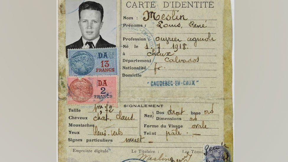 A close-up of a fake German-issued World War Two identity card issued for Lonnie Moseley, giving his name as Louis Rene Meslin. It shows his photo top left and is a yellow/sepia coloured rectangular piece of card. Typewritten on it are lists of required information such as first name and surname, his profession, height, eye colour etc. These details are filled in by hand. Official stamps are along its bottom quarter. 