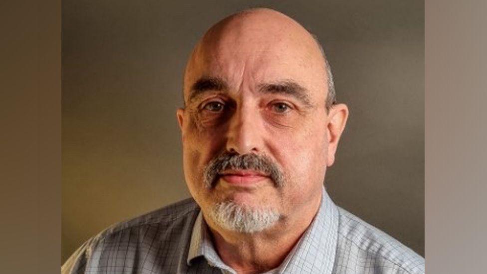 Professional headshot of Keith Sandle. He is wearing a white shirt has greying hair and a grey beard.