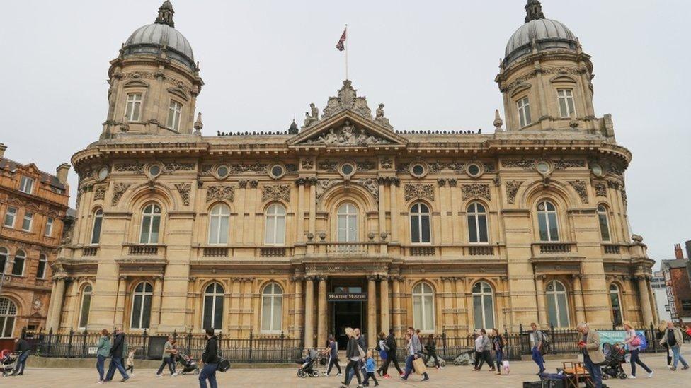 View of the outside of the maritime museum, which is a two-storey Victorian building of yellow stone with two domes at the top. People are walking in front of it as they go about their daily business.