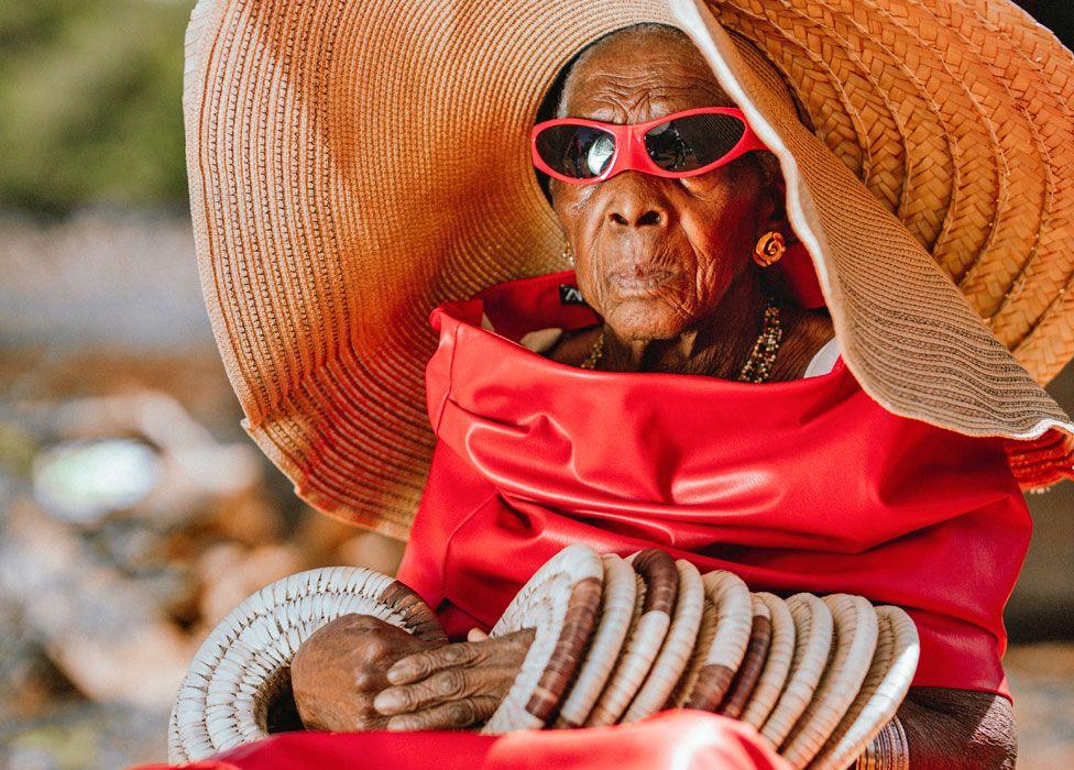 A seated Margret Chola looks serious as she wears a giant straw hat with bangles that look like straw mat up both her arms. She is wearing red sunglasses and what looks like a red leather dress