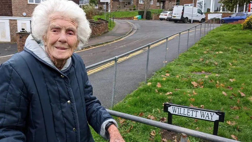 Joan stands on the street in front of a metal handrail and a sign which reads "Willett Way". She is looking at the camera and smiling, and is wearing a dark blue coat with the cuffs turned slightly up.